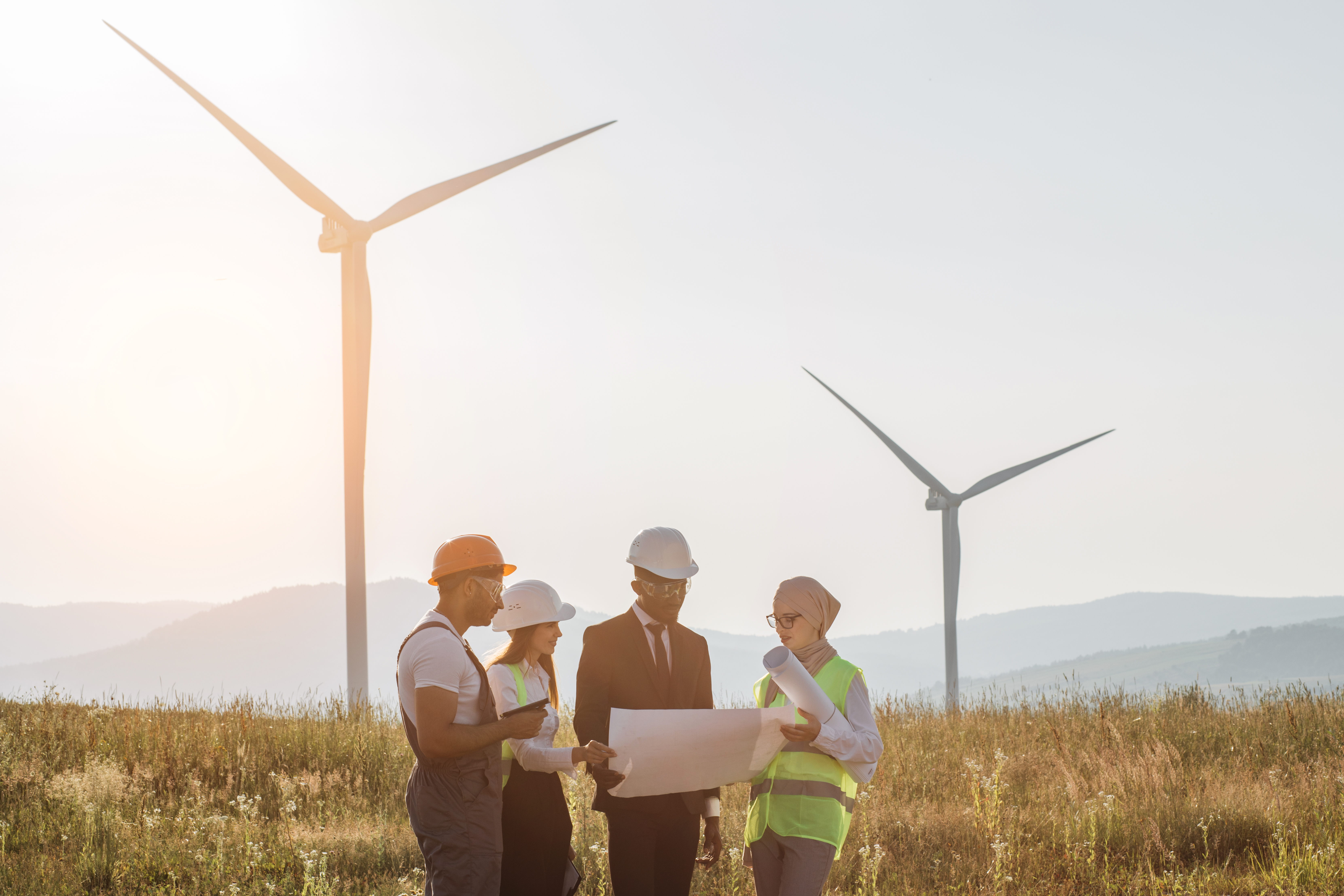 People stand in front of wind turbine blades on the ground and a working wind turbine.